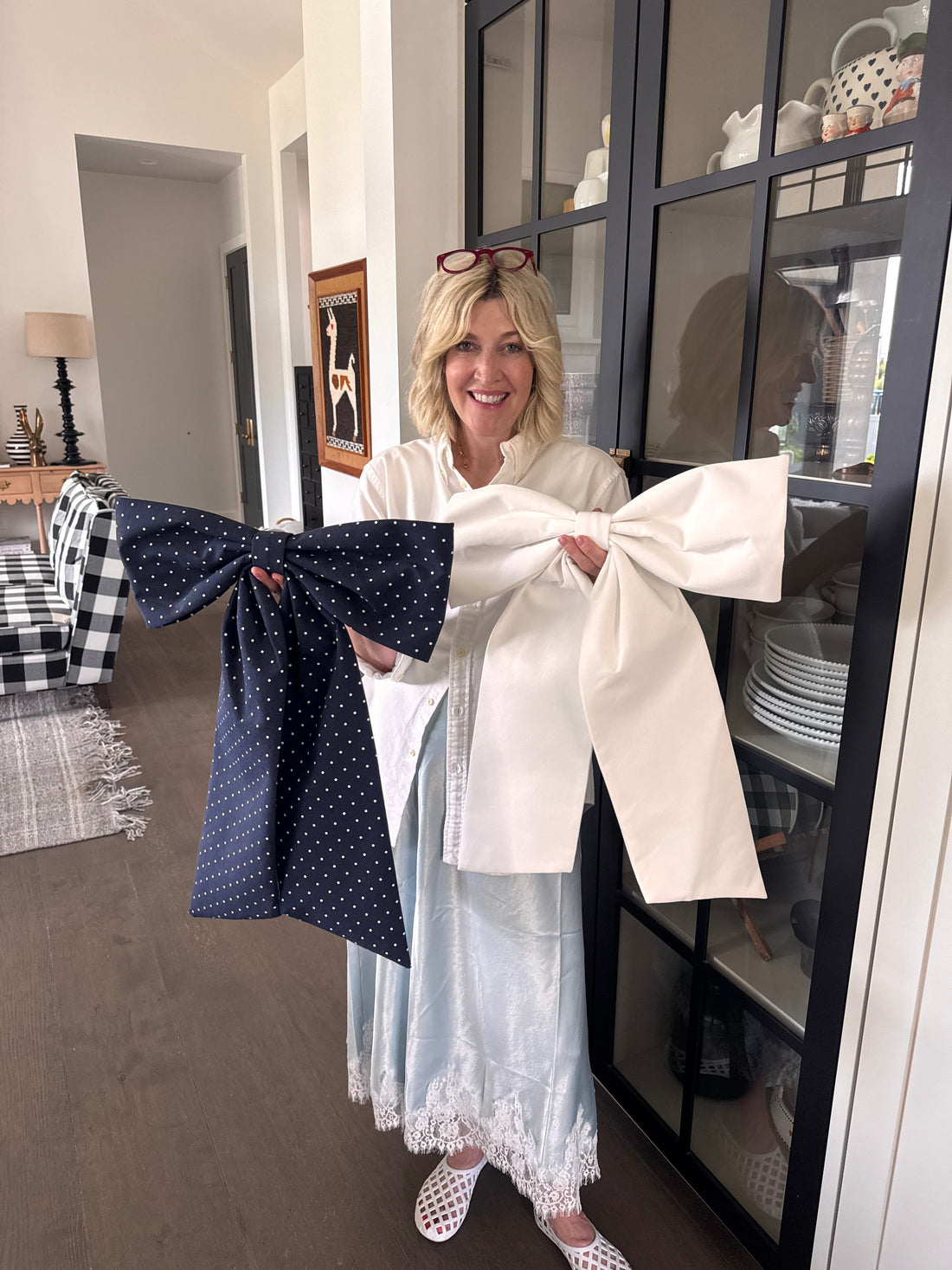 Jeneil smiles at the camera while standing in her living room, holding a white door bow and a navy polka dot door bow