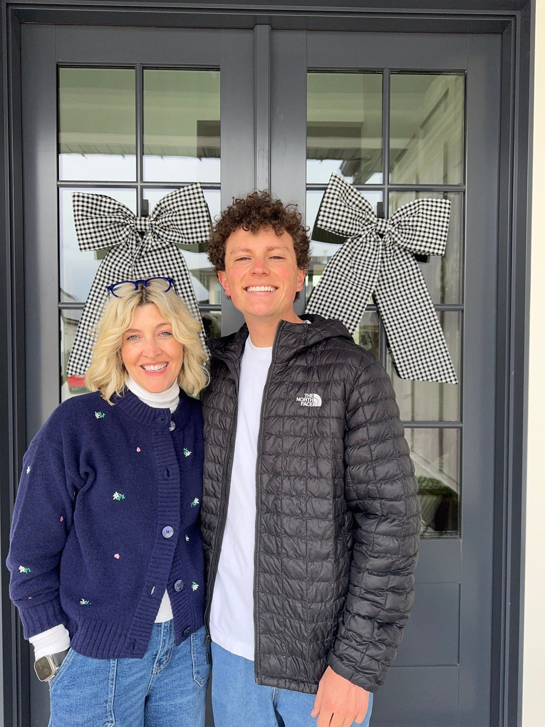 Jake and Jeneil stand in front of double doors with black gingham bows and smile