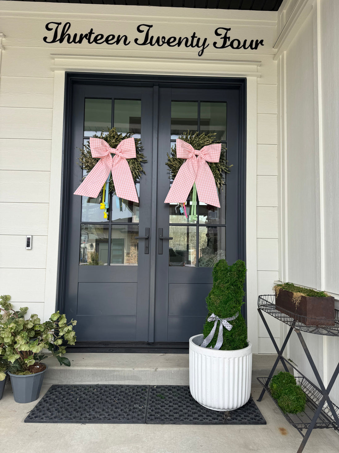Two pink holiday bows on a set of double front doors, with a potted bunny shaped bush hedge in front