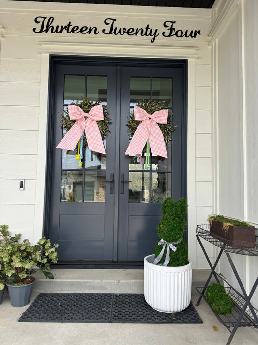 Two pink holiday bows on a set of double front doors, with a potted bunny shaped bush hedge in front