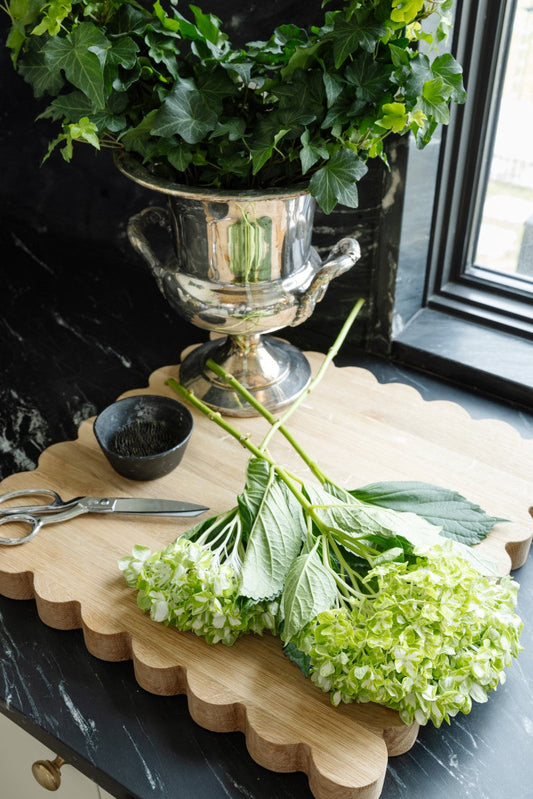 Scalloped oak cutting board with green flowers, a pair of silver scissors, and a copper plant holder sitting on it