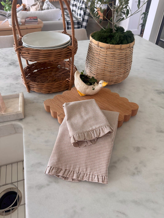 A cream gingham dish cloth and gingham hand towel on a white countertop, staged with an oak trivet, a duck shaped plant holder, and a plant in a wicker basket