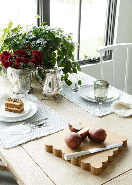 A table set with greenery, a scalloped oak cutting board with pears, and ruffled placemats set with white dishes and carrot cake