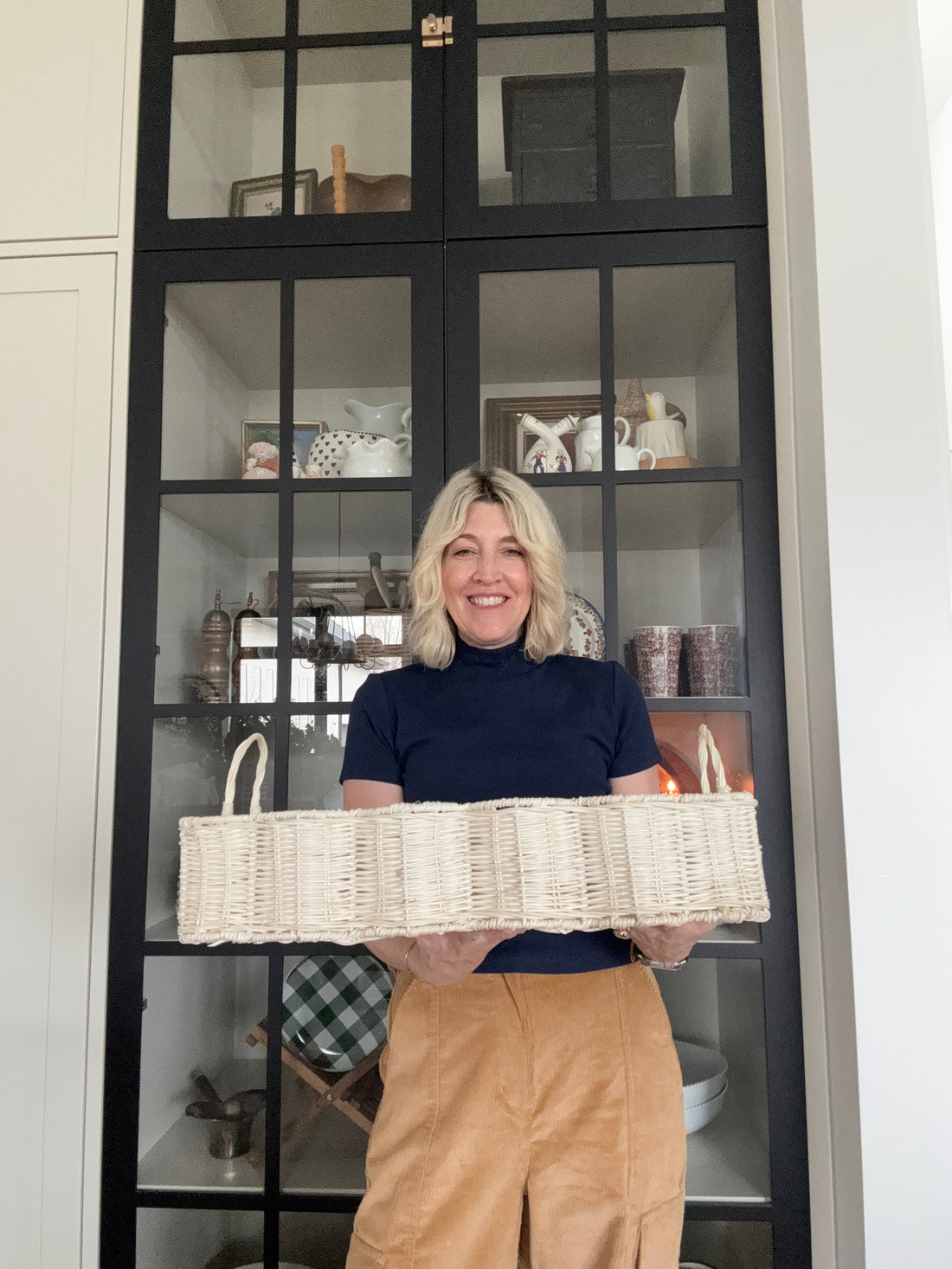 Jeneil stands in front of a navy blue and glass cabinet holding a scalloped wicker basket
