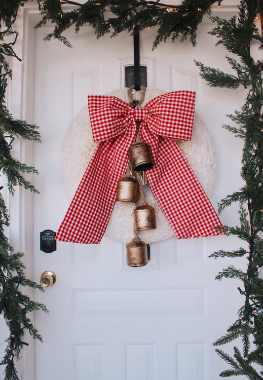 Red gingham hung with white bow on front door with bells hung on the wreath and green garland hung on the border of the door